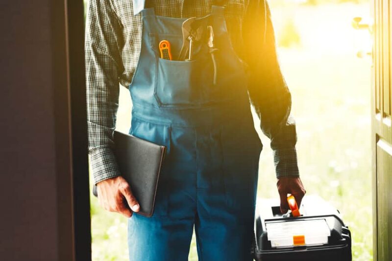 a plumber entering a home while carrying paperwork and a toolbox