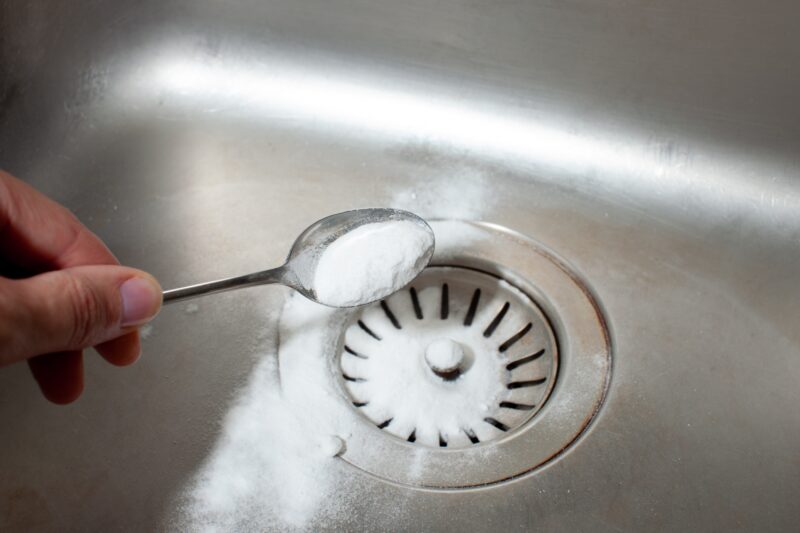 a hand dumping a spoonful of baking soda into a drain as part of a DIY drain clog removal