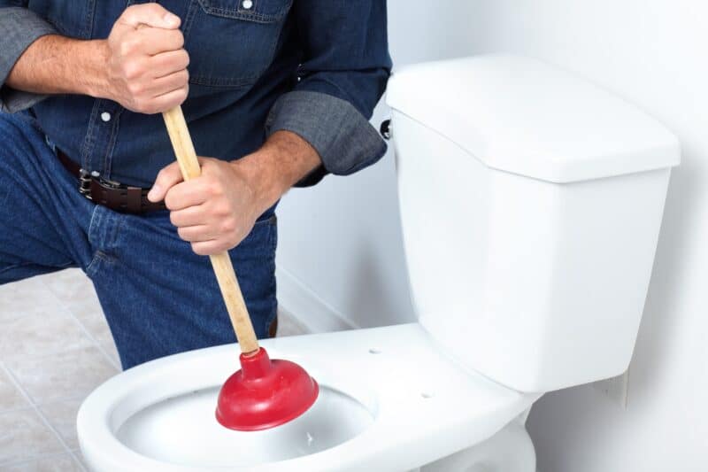 a professional plumber kneeling down to use a plunger to clear a toilet clog